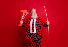 Photo of crazy cupid mature handsome guy valentine day hold bow arrow, ready to shoot loving couple wear hearts pattern stylish suit tie trousers blazer isolated red color background